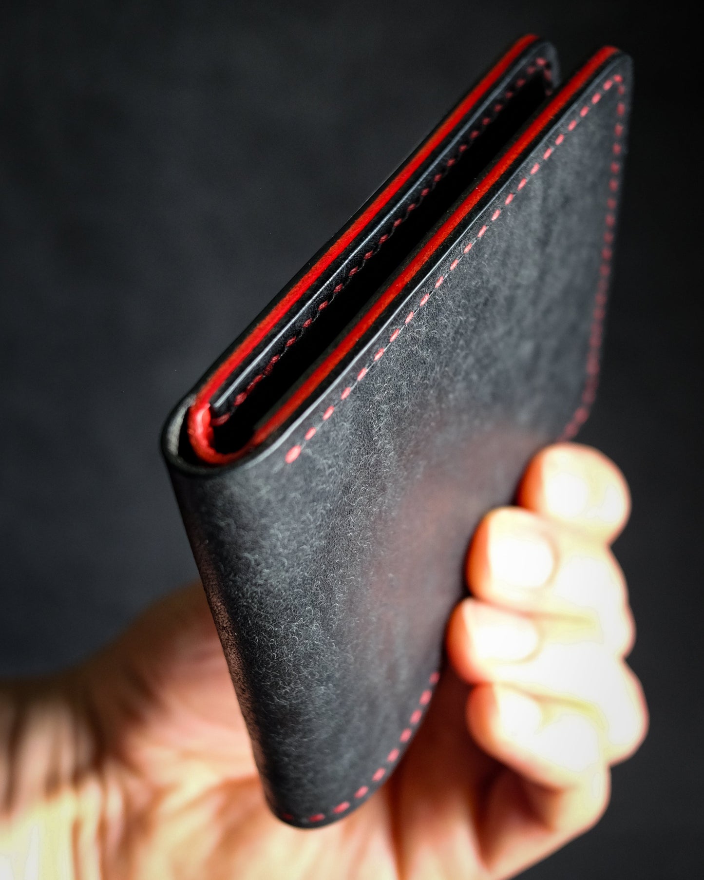 Hand holding a black leather wallet with red stitching against a dark background